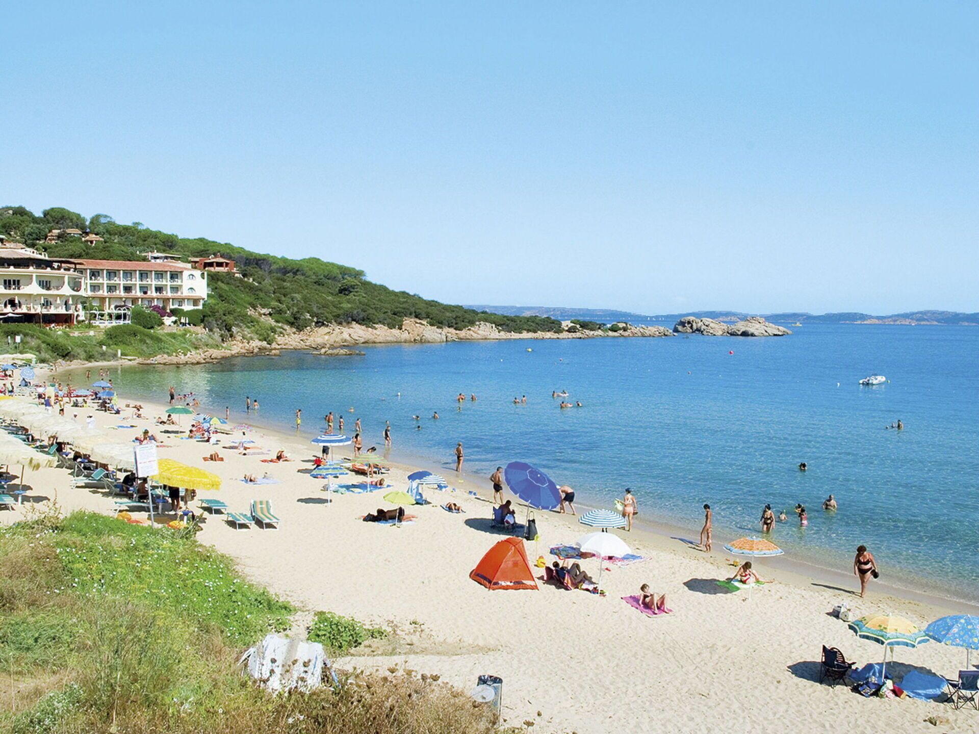 A beach with people swimming and sunbathing, umbrellas, and a building in the background.