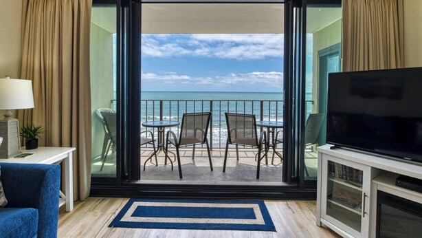 A room with a balcony overlooking the ocean, furnished with a TV, a small table, and two chairs. - Oceanfront Oasis - 2 Bedroom, 2 Bath Beautifully Renovated Oceanfront Condo (Garden City Beach)