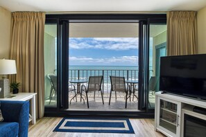 A room with a balcony overlooking the ocean, furnished with a TV, a small table, and two chairs.