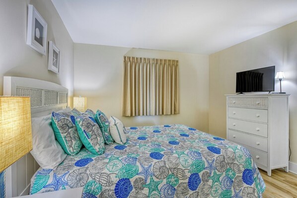 Une chambre à coucher avec un lit, une commode, un téléviseur et une fenêtre avec des rideaux.