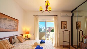 Une chambre d’hôtel comprenant un lit, une table de chevet, une lampe, un tableau, une chaise, une table, un vase avec des fleurs et un appareil de climatisation.
