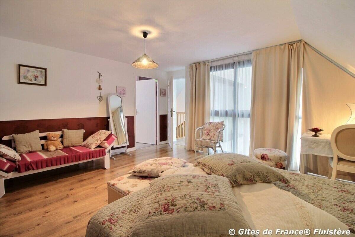 A bedroom with a wooden floor, a bed with floral bedding, a sofa with a teddy bear, a chair, and a table with a lamp.