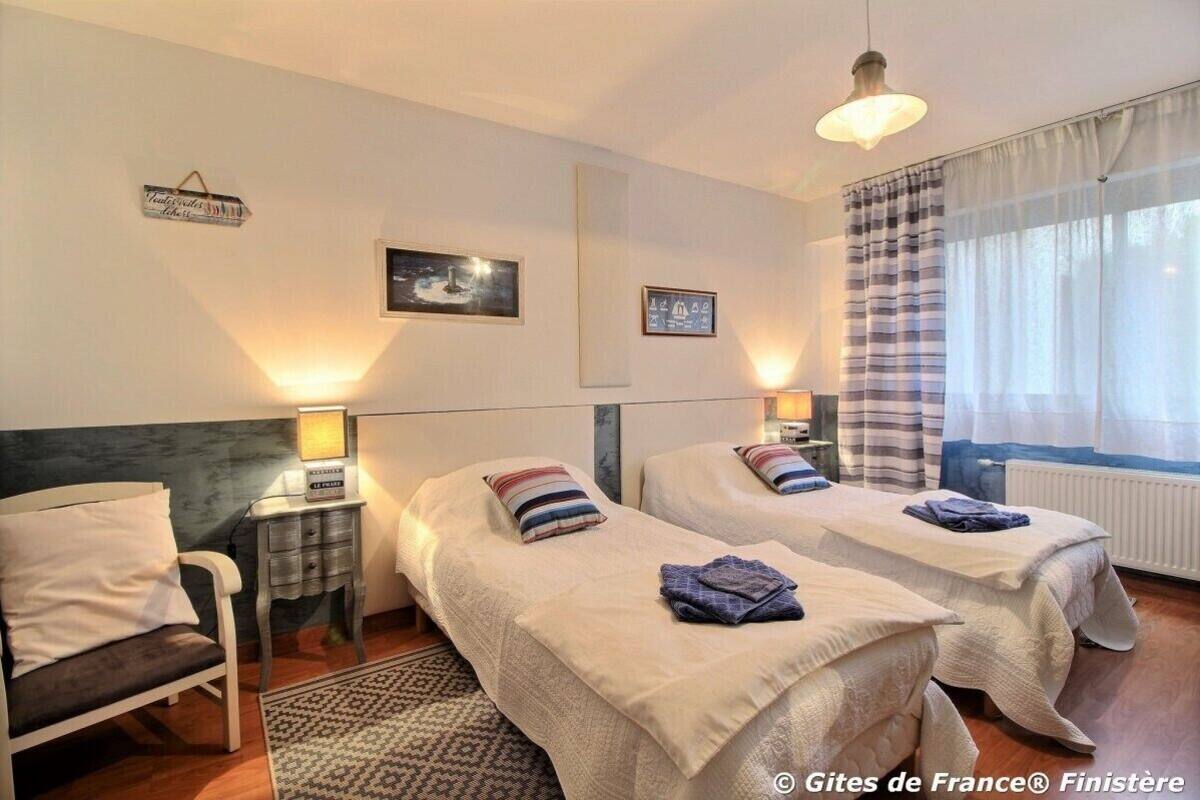 A hotel room with two beds, a small table with a lamp, a chair, and a window with curtains.
