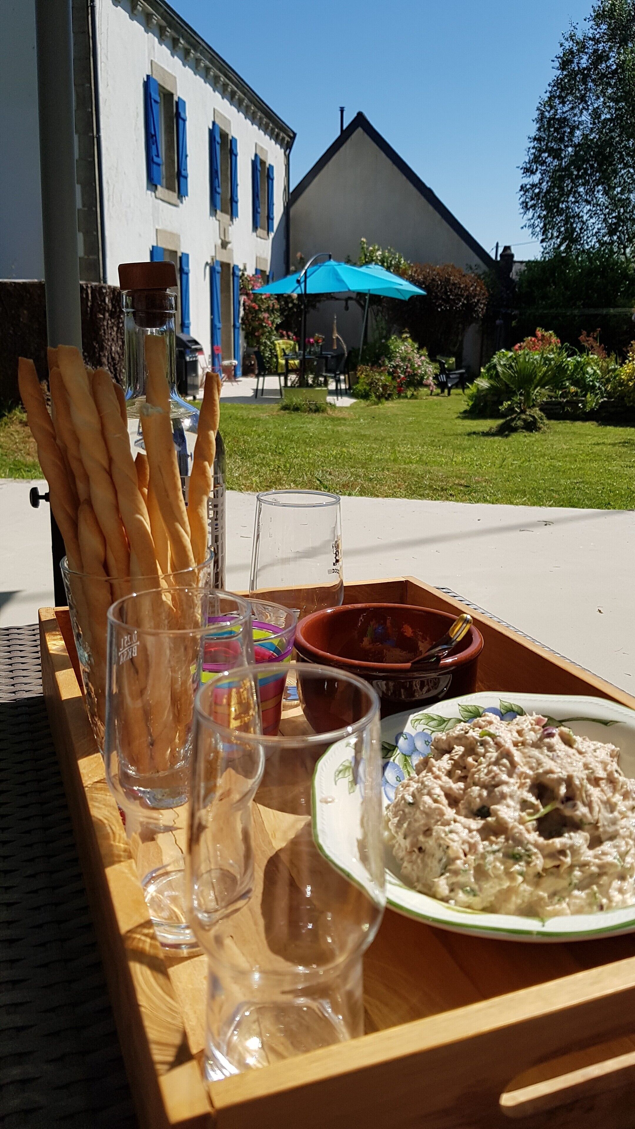 Una mesa con hogazas de pan, un plato de ensalada y un vaso de agua, colocada al aire libre con un edificio y un jardín al fondo.