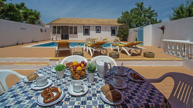 Un espace extérieur avec une piscine, des chaises longues, une table à manger où sont servis les articles du petit-déjeuner, et un bâtiment en arrière-plan.