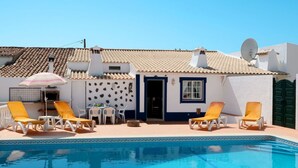 Un espace aménagé au bord de la piscine, avec des chaises longues, une table et une porte donnant accès à un bâtiment doté d’un toit en tuiles.