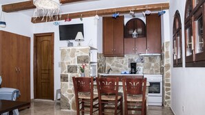 A kitchen with wooden cabinets, a stone wall, a dining table with chairs, and a TV mounted above the cabinets. - Bella Vita Style rooms & apartments-Incanto (Pitsidia)