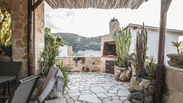 A stone-tiled patio with a table and chairs, surrounded by cacti and plants, overlooking a body of water and hills. - Bella Vita Style rooms & apartments-Incanto (Pitsidia)
