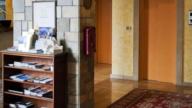 Ένας χώρος υποδοχής με έναν τοίχο από πέτρα, κορνιζάκια με φωτογραφίες, μια βιβλιοθήκη, ένα κόκκινο χαλί με σχέδια και μια πόρτα.