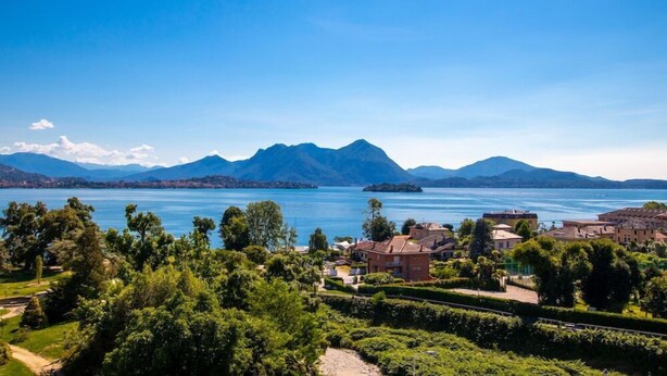 A lakeside view with mountains in the background and a residential area with houses and greenery in the foreground. - Alpi (Baveno)