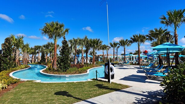 A pool area with a curved design, surrounded by palm trees and lounge chairs under blue umbrellas. - Game Room • Private Pool & Spa • Lazy River • Near Golf • Gated • Near Disney (Kissimmee)