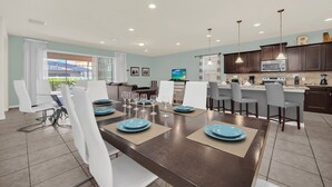 A modern dining area with a dark wooden table set for six, surrounded by white chairs. A kitchen with dark cabinets and a microwave is visible in the background. - Game Room • Private Pool & Spa • Lazy River • Near Golf • Gated • Near Disney (Kissimmee)