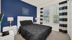 A bedroom with a black bedspread, white headboard, and a decorative wall piece. There are two bedside tables with lamps, a window with striped curtains, and a view of greenery outside. - Game Room • Private Pool & Spa • Lazy River • Near Golf • Gated • Near Disney (Kissimmee)