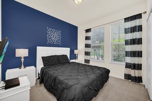A bedroom with a black bedspread, white headboard, and a decorative wall piece. There are two bedside tables with lamps, a window with striped curtains, and a view of greenery outside.