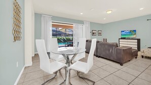 A modern living room with a glass dining table, white chairs, a brown leather sofa, and a TV. - Game Room • Private Pool & Spa • Lazy River • Near Golf • Gated • Near Disney (Kissimmee)