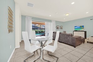 A modern living room with a glass dining table, white chairs, a brown leather sofa, and a TV.