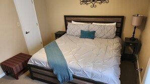 A neatly made bed with a wooden frame, a bedside table with a lamp, and a wall-mounted decorative piece. - Great Home; Close to Everywhere. (Oak Grove)