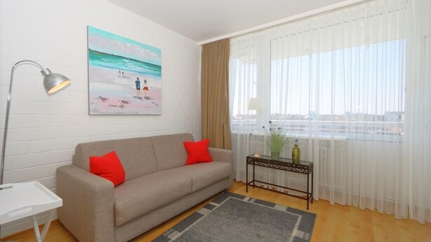 Un salon avec un canapé beige, des coussins rouges, un tableau représentant une scène de plage, une lampe, une table avec un vase et des rideaux en voile.