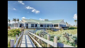 Een strandhuis met een houten looppad dat naar een woning leidt, met een groen dak en meerdere balkons.