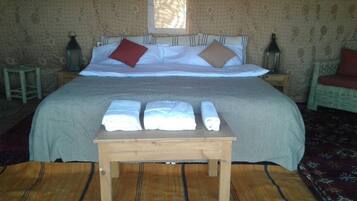 A neatly made bed with pillows, a small wooden table with towels, and a window with curtains.