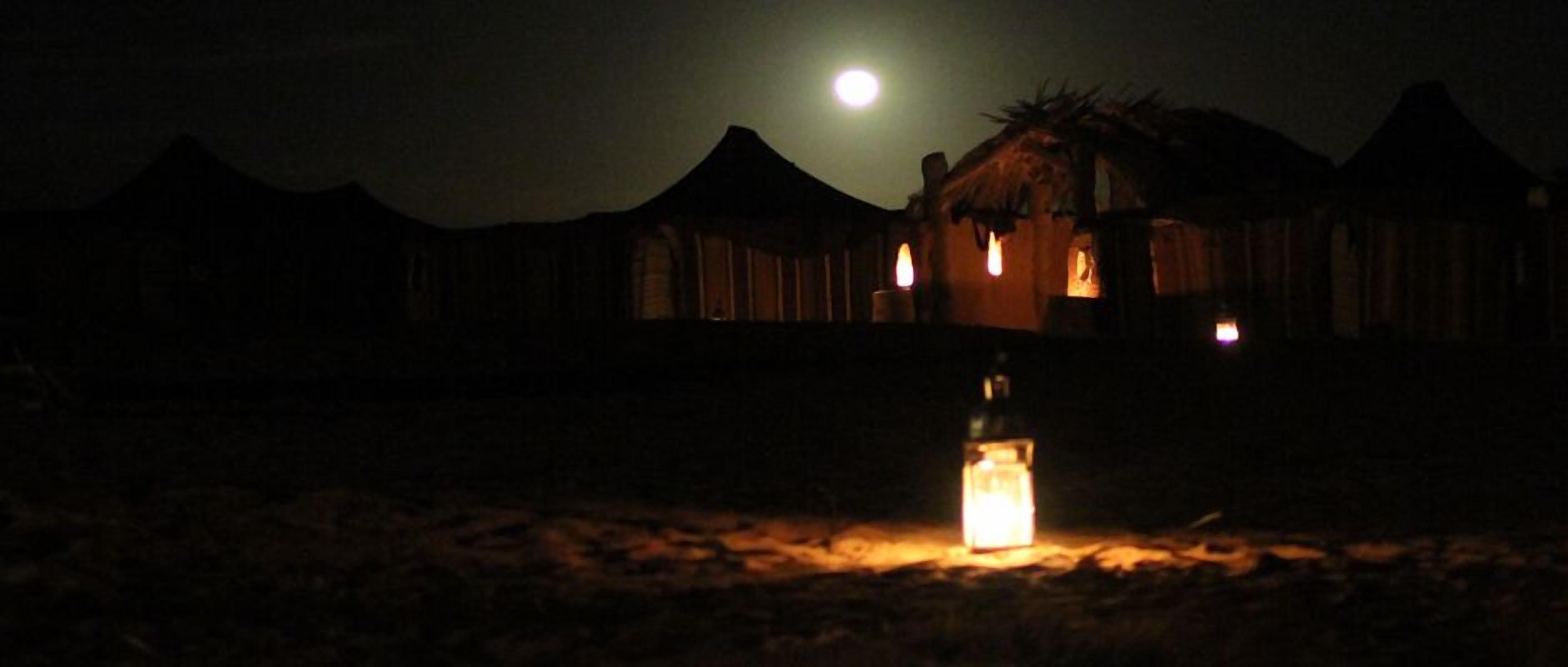 A night scene with a lit candle in the foreground, a moon in the sky, and a building with a thatched roof in the background.