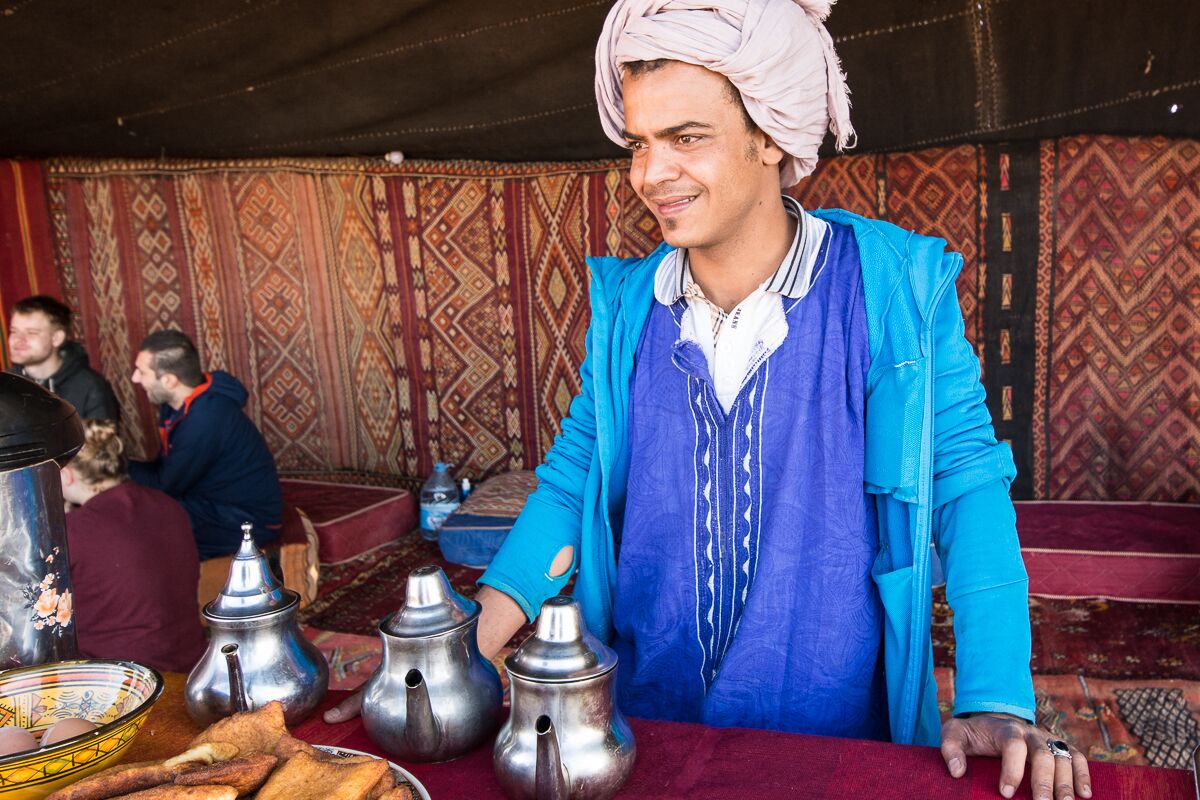 A person in traditional attire standing behind a table with teapots and a bowl of food.