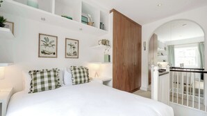 A bedroom with a large bed, white walls, and a wooden wardrobe. There are shelves with decorative items and a window with curtains.