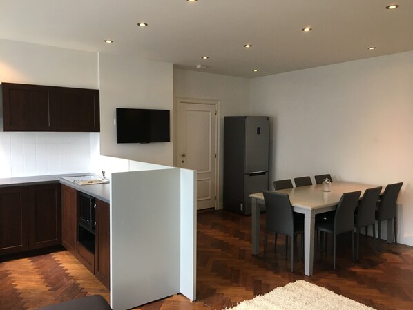A modern kitchen with a dining area, wooden flooring, and a flat-screen TV mounted on the wall.