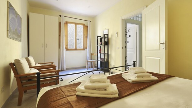 Un dormitorio con una cama, un banco de madera, una ventana, una puerta y una estantería con toallas.