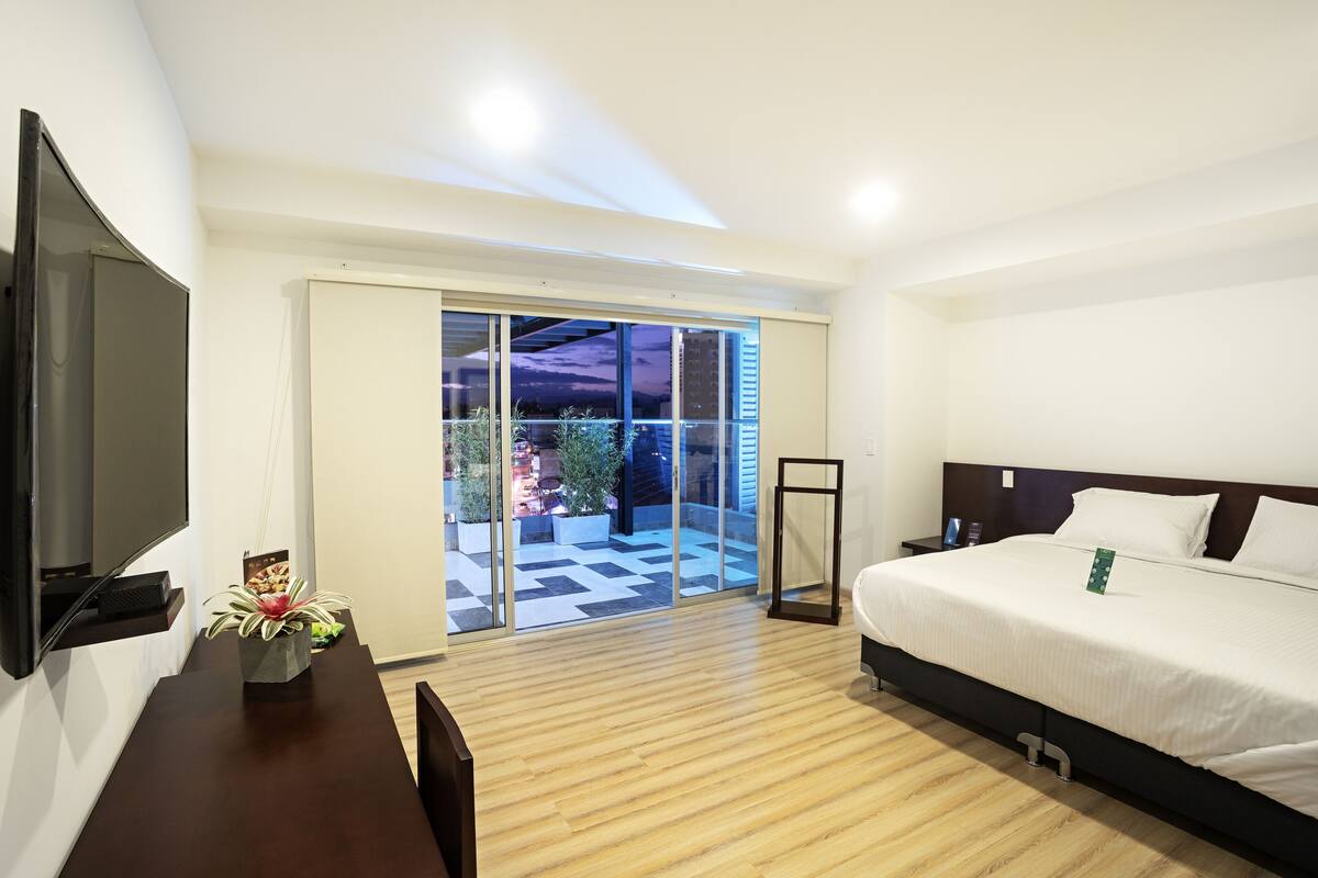 A modern hotel room with a large bed, a flat-screen TV mounted on the wall, a wooden floor, and a sliding glass door leading to an outdoor area with plants.