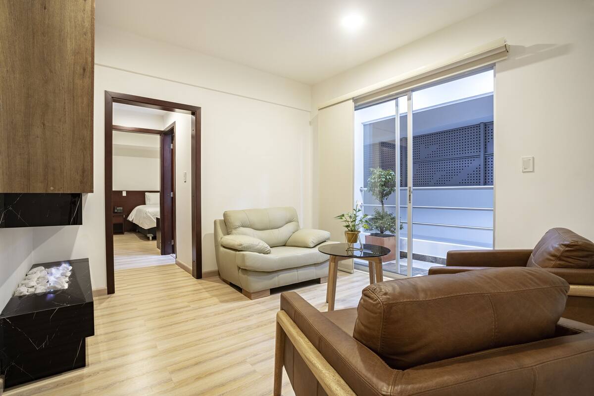 A modern living room with a sofa, a coffee table, and a TV. There is a door leading to another room and a large window with a view.