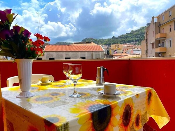 A table with a sunflower tablecloth, a white vase with flowers, two wine glasses, a coffee pot, and a cup on a balcony overlooking buildings and hills. - Aurora, in the center of Tropea 350m from the sea with air conditioning (TROPEA)