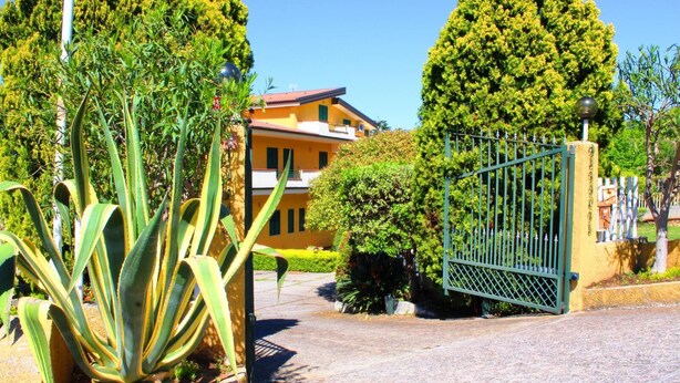 A gated entrance with a paved driveway leading to a yellow building with a red roof, surrounded by lush greenery and a tall tree. - Sole apartment with sea view and free wi fi (n ° 3) (Santa Domenica)