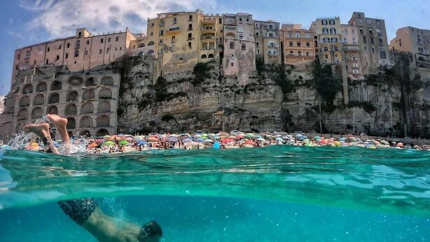 A swimmer in clear water with a rocky cliff and colorful buildings in the background. - Gaia Apartment with Sea View, 350m from the white beach of Tropea (TROPEA)