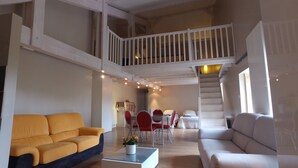 Ein großzügiger Wohnbereich mit gelbem Sofa, weißem Sofa und einem Esstisch mit roten Stühlen. Eine Treppe führt zu einer oberen Ebene.