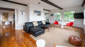 Ein modernes Wohnzimmer mit einer schwarzen Eckcouch, einem Essbereich mit Holztisch und -stühlen sowie einem großen Fenster, das für natürliches Licht sorgt.