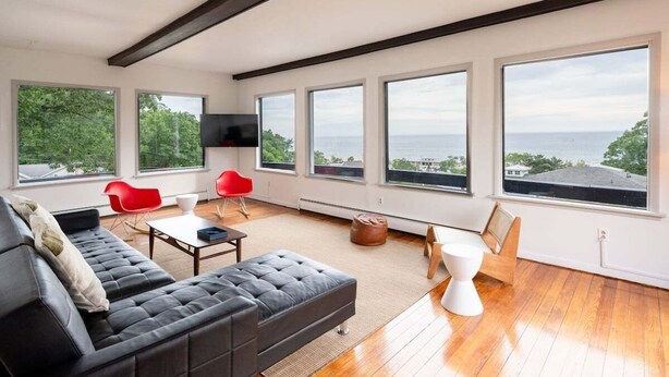 Ein geräumiges Wohnzimmer mit einer großen schwarzen Couch, roten Sesseln, einem weißen Couchtisch und einem Holzespeisetisch. Große Fenster bieten Blick auf das Meer und die grüne Landschaft.