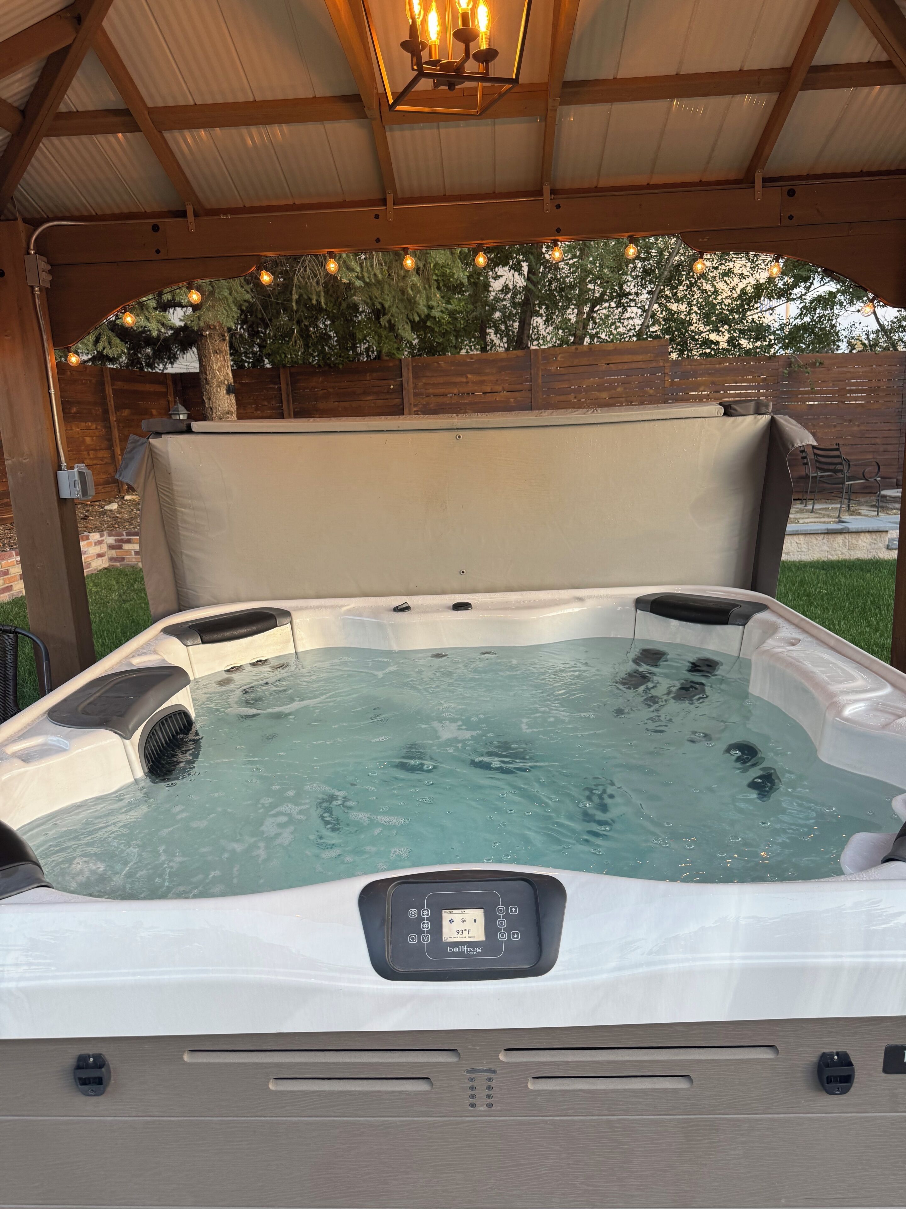 Un jacuzzi con panel de control, rodeado por una terraza de madera y una zona cubierta con luces colgantes.