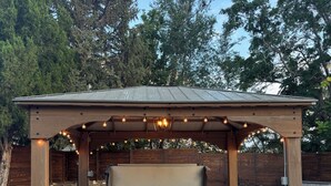 A hot tub under a wooden pergola with string lights, surrounded by a brick patio and a grassy area.