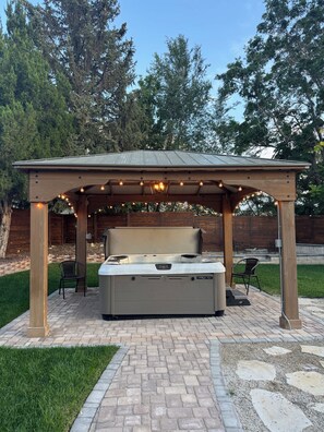 Un bain à remous sous une pergola en bois, illuminé de guirlandes lumineuses, entouré d’une terrasse en briques et d’une pelouse.