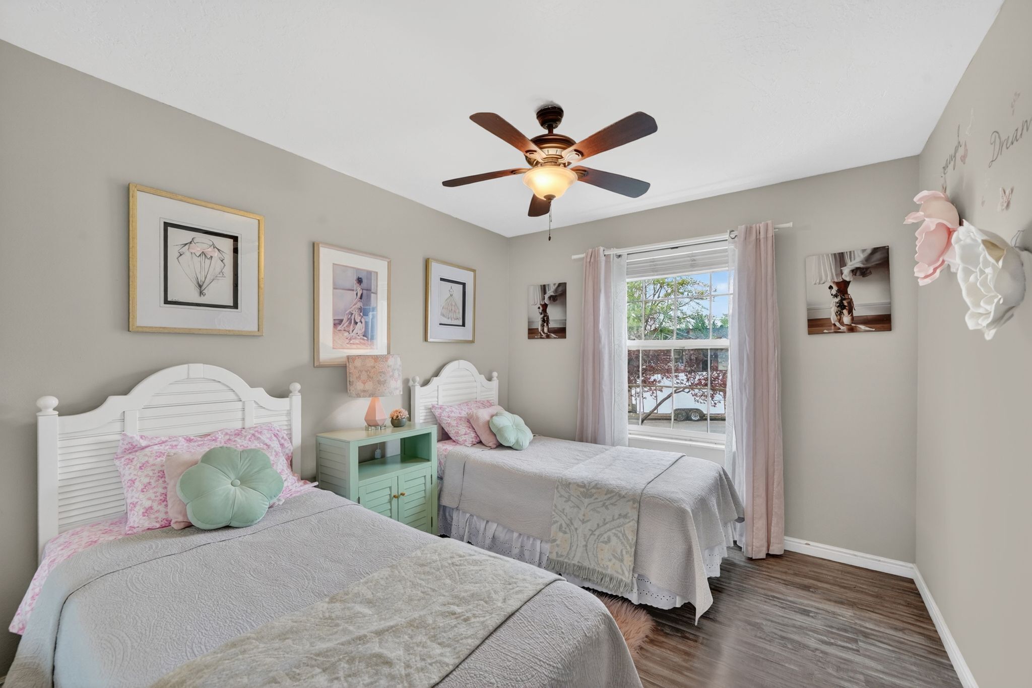 A bedroom with two beds, a ceiling fan, framed artwork, and a window with curtains.
