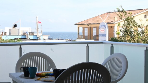 A balcony with a table and chairs, overlooking the sea and a building with a sign. - M033 - Marcelli, three-room apartment in the center facing the sea (Marcelli)