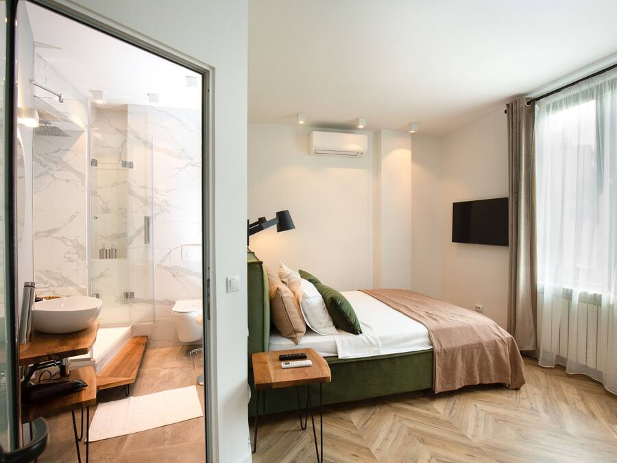 A modern hotel room with a glass shower enclosure, a bed with a green headboard, a bedside table with a remote control, and a large window with sheer curtains.