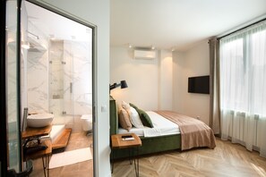 A modern hotel room with a glass shower enclosure, a bed with a green headboard, a bedside table with a remote control, and a large window with sheer curtains. - Metropole Apartments Royal Residence (Bucharest)