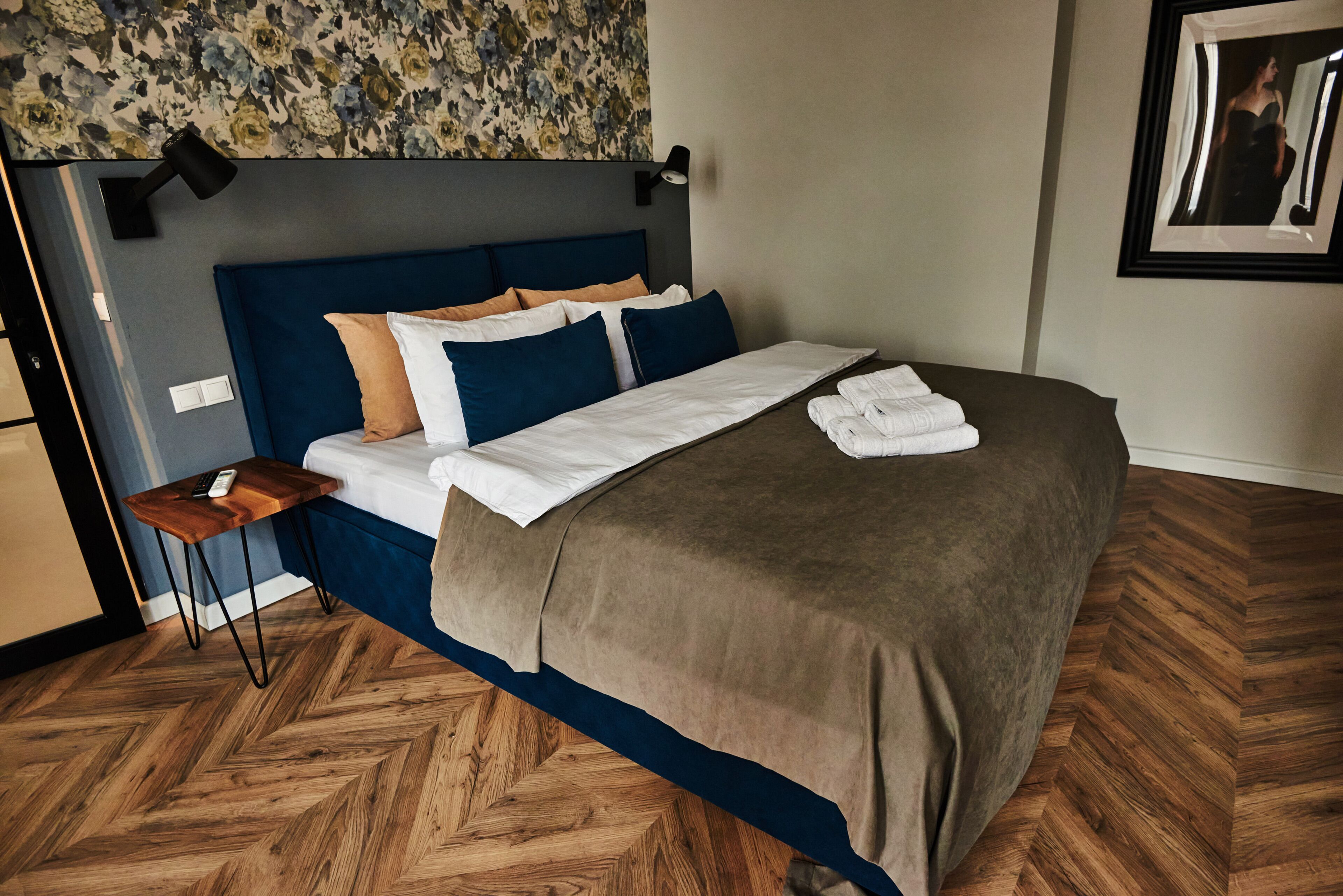 A neatly made bed with a blue headboard, a wooden bedside table with a remote control, and a patterned wallpaper above the bed.