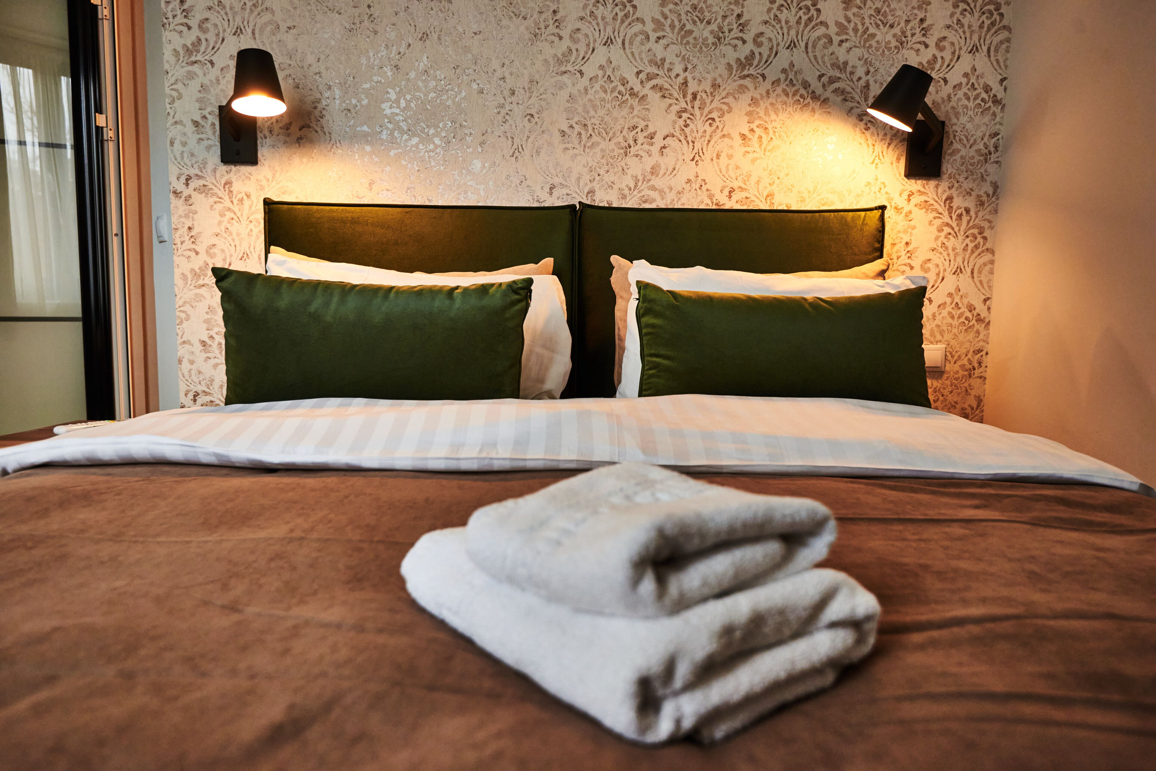 A neatly made bed with green pillows, a folded white towel, and two wall-mounted lamps.