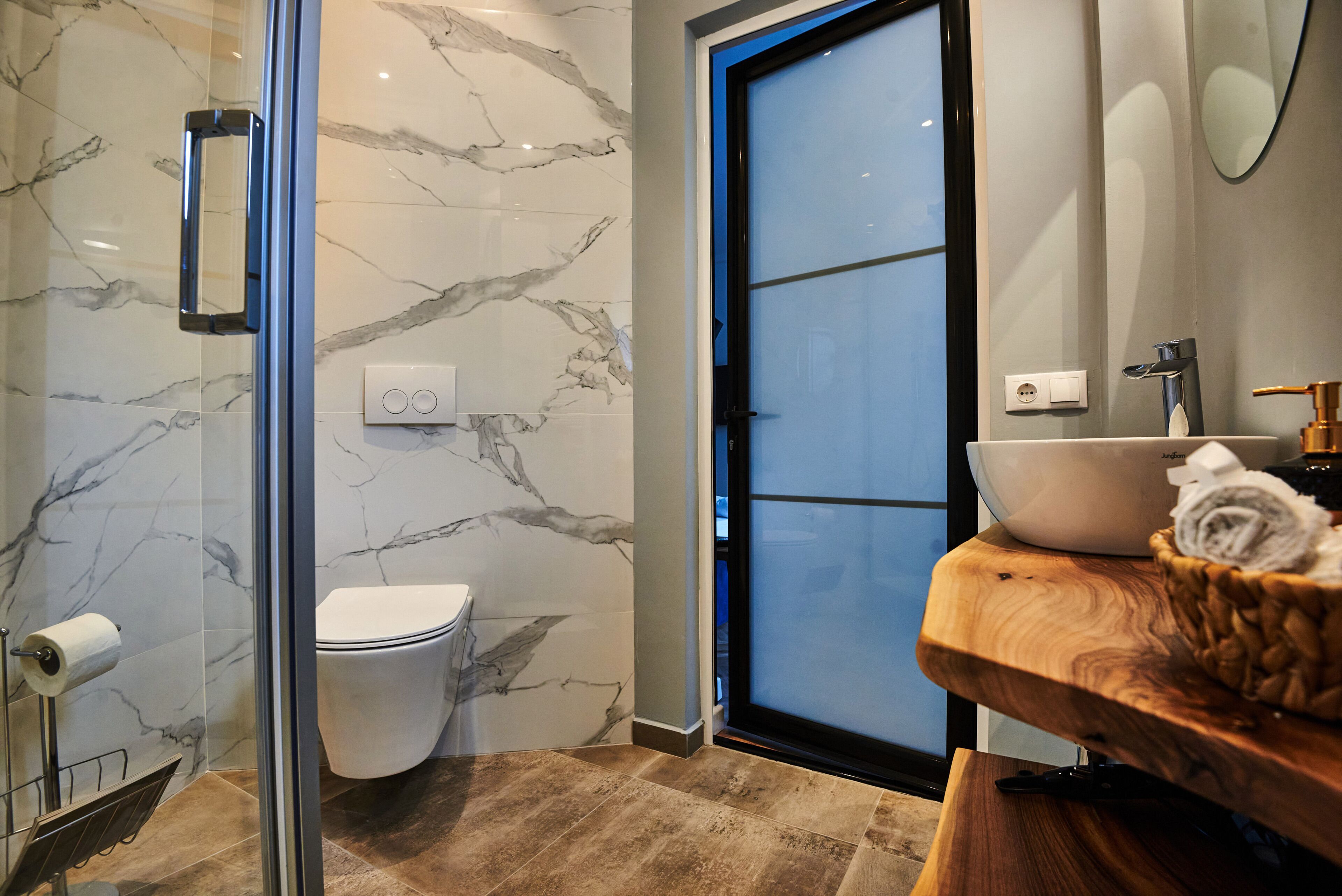 Ein modernes Badezimmer mit einer Duschkabine aus Glas, einer Wand mit Marmorplatten, einem Holzwaschtisch und einer Toilette.