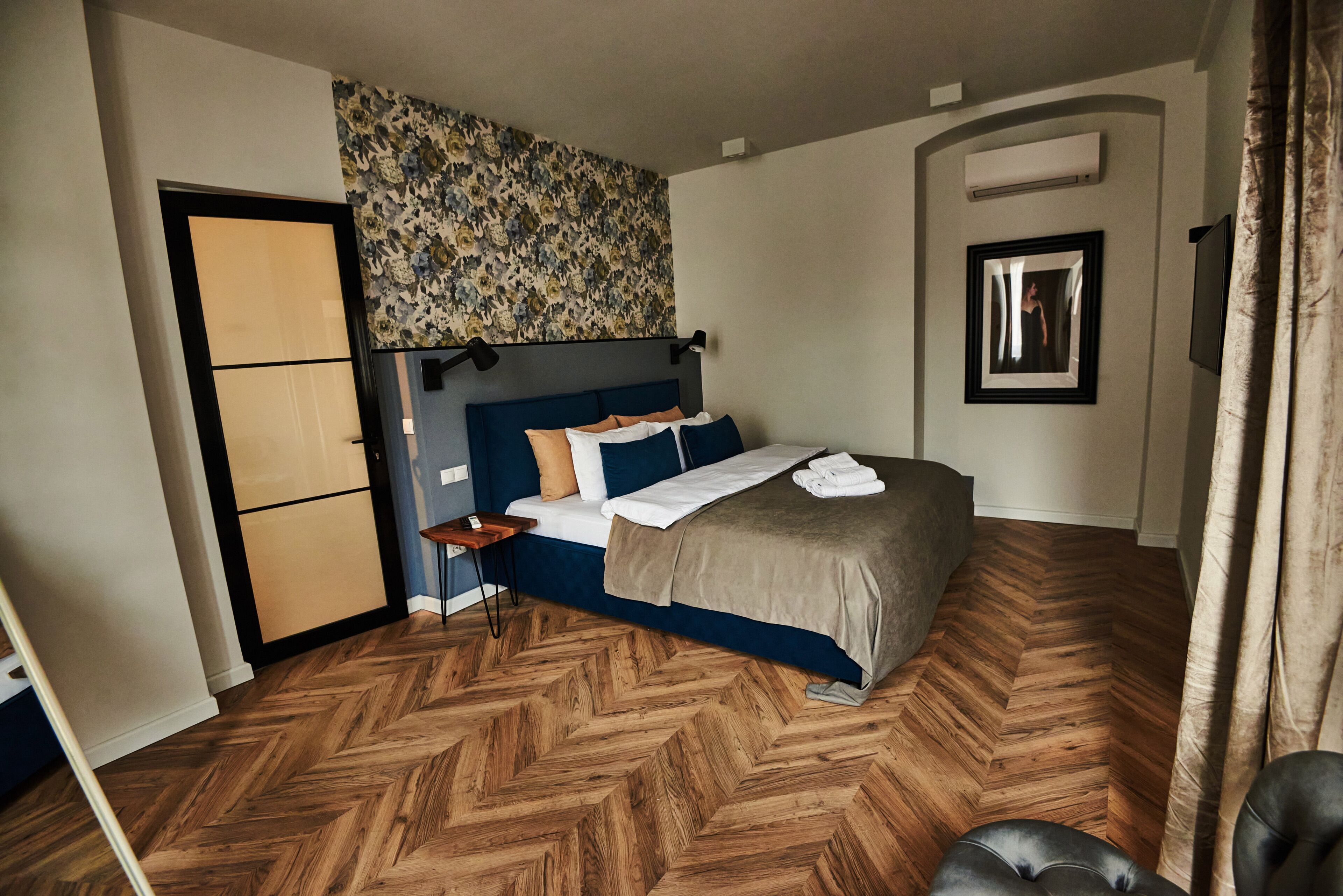 A bedroom with a bed, a nightstand, a mirror, and a herringbone-patterned wooden floor.