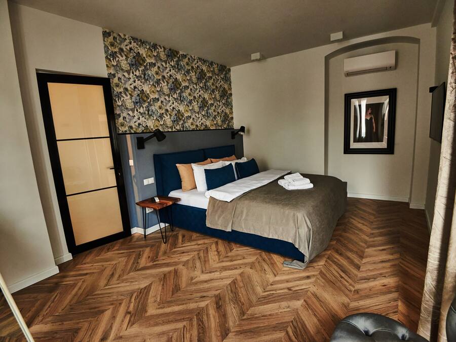 A bedroom with a bed, a nightstand, a mirror, and a herringbone-patterned wooden floor.