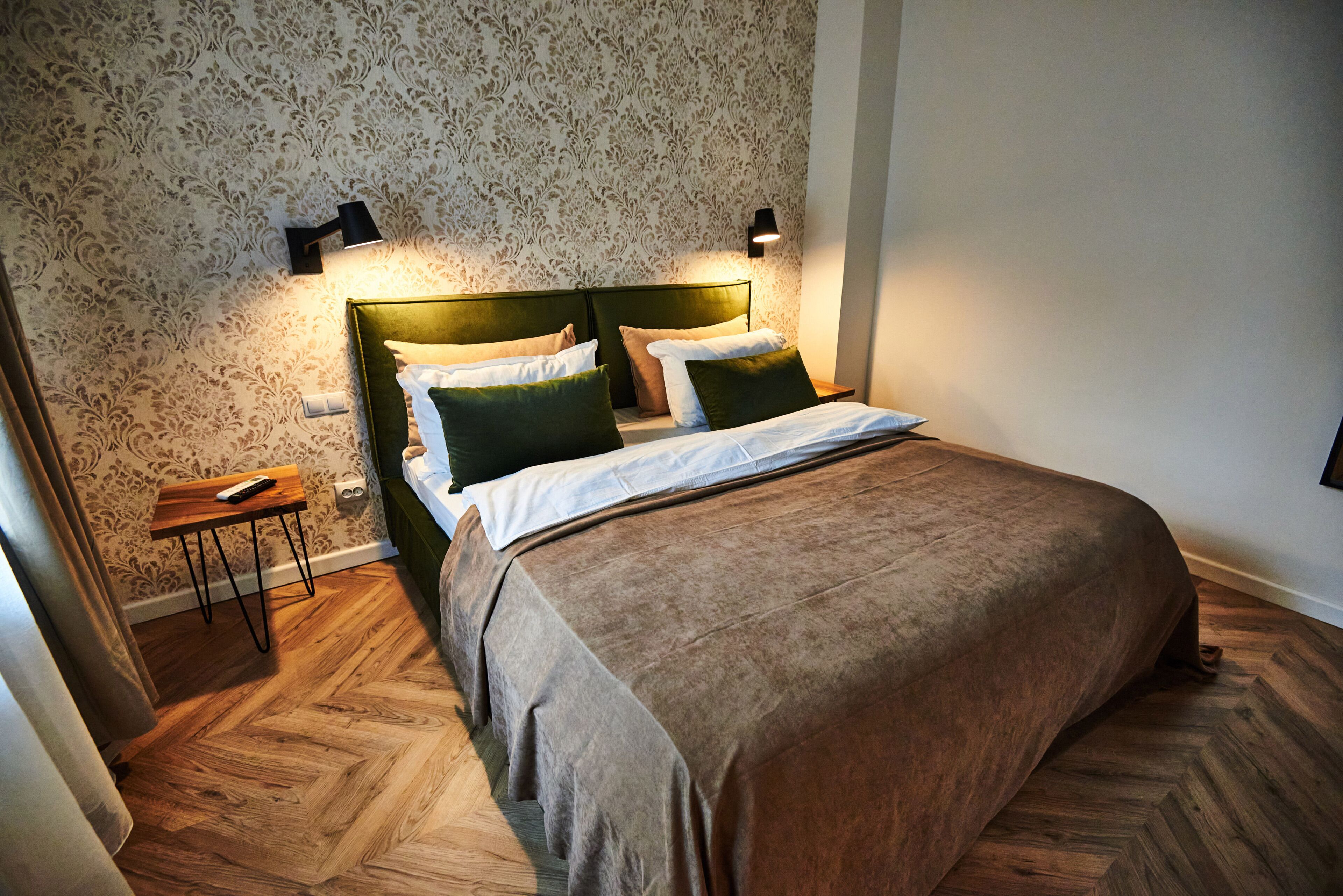 Een net opgemaakt bed met een groen hoofdbord, twee bedlampjes en een houten nachtkastje met een afstandsbediening.